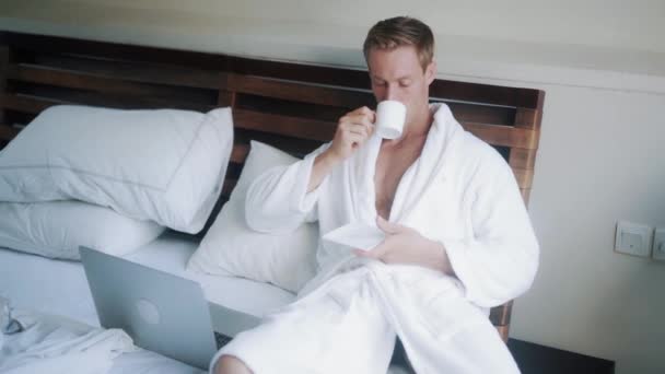 young man in bathrobe drinks coffee in bed near laptop - Video