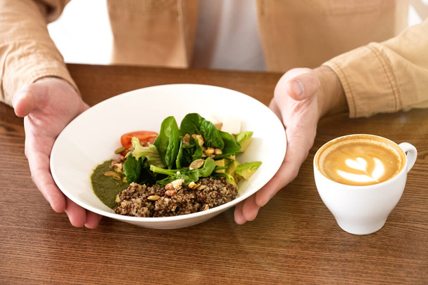 άνθρωπος κρατώντας πιάτο της σαλάτας και με τα δύο χέρια πριν από την έναρξη του γεύματος με ζεστό καφέ στο καφέ κατά τη διάρκεια του διαλείμματος. - Φωτογραφία, εικόνα