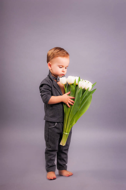 Un pequeño caballero con un ramo de tulipanes en el estudio sobre un fondo gris en honor a las vacaciones de las mujeres el 8 de marzo y una tarjeta de felicitación en forma de todas las madres, abuelas, niñas, niñas
 - Foto, Imagen