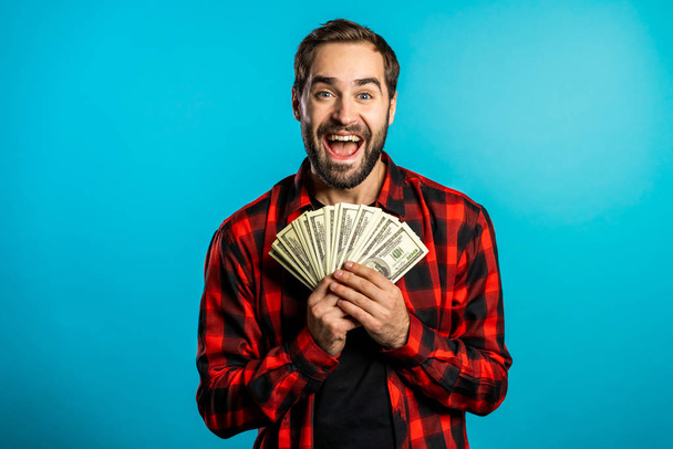 Satisfied happy excited man showing money - U.S. currency dollars banknotes on blue wall. Symbol of success, gain, victory. - Foto, Bild