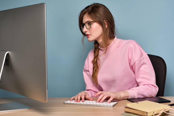 A girl with a braid in a pink sweater and glasses sits at a table and looks at the monitor, typing on a keyboard. - Photo, image