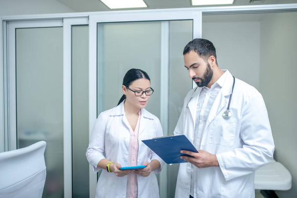 Dark-haired male and female doctors standing in the room together discussing schedule - Foto, imagen