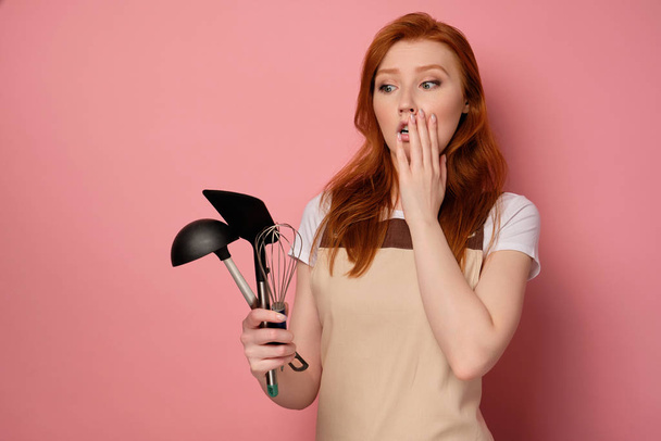 A red-haired girl in a beige apron and a T-shirt stands on a pink background, looks with horror at the kitchen appliances in hand - Photo, image