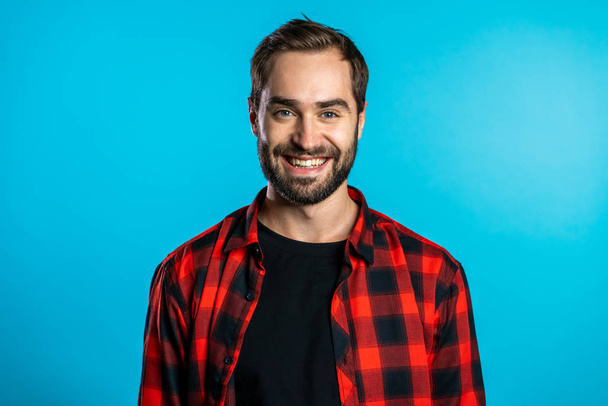 Handsome european man with trendy beard in red plaid shirt on blue studio background. Cheerful guy smiling and looking to camera. - Foto, Imagen