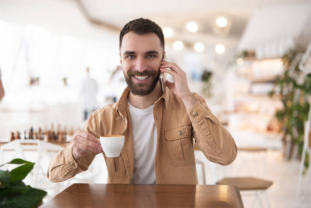 Joven hombre guapo bebe café en la cafetería para el almuerzo hablando en su teléfono inteligente, buscando satisfecho, concepto multitarea
. - Foto, Imagen