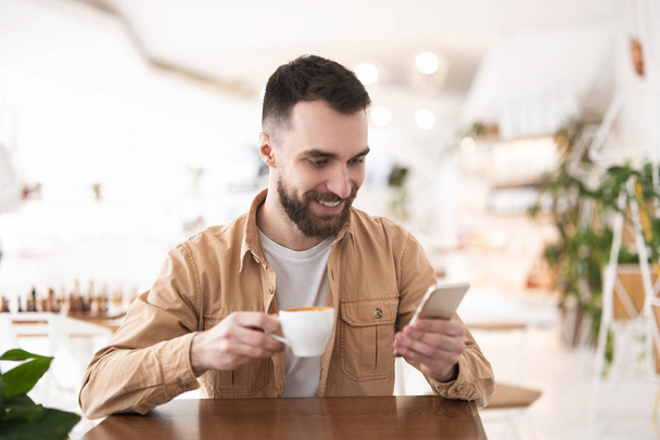 Young bearded man checking news in his smartphone while having his morning coffee at the cafe, everyday routine. - Photo, image