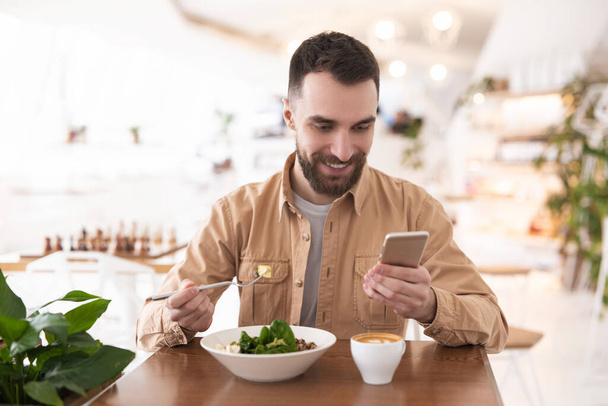 Young bearded smiling man drinks his hot coffee and eats salad for lunch while checking news in his smartphone during break , multitasking concept. - Fotó, kép