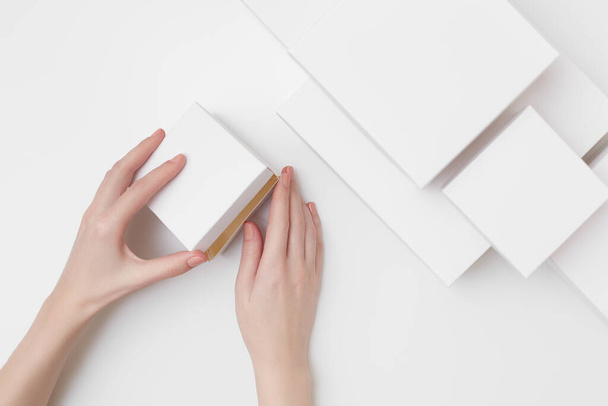 Макет білої подарункової коробки в жіночих руках на білому тлі в оточенні багатьох різних розмірів білих коробок
 - Фото, зображення