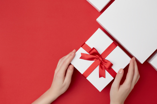 Womens hands holding white present box with red ribbon surrounded with white boxes on the red background - Photo, Image
