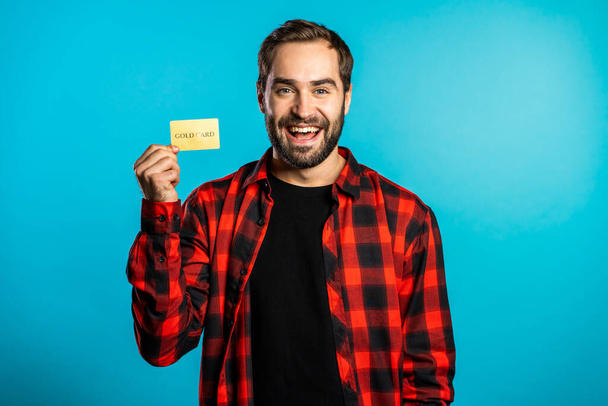 Successful man in plaid shirt showing unlimited gold credit card on blue studio background. Student, money concept - Foto, Imagem