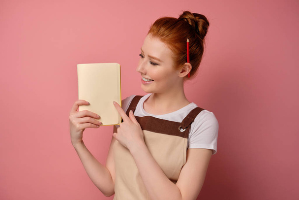 Egy cuki vörös hajú lány, magas zsemlével bézs kötényben, rózsaszín háttéren áll, és egy notebook oldalát mutatja.. - Fotó, kép