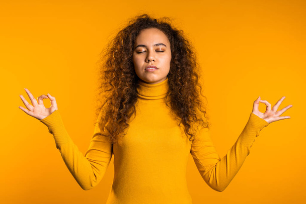 Positive curly girl relaxing, meditating. Woman calms down, breathes deeply with mudra om on yellow studio background. Young curly woman portrait. - Fotografie, Obrázek