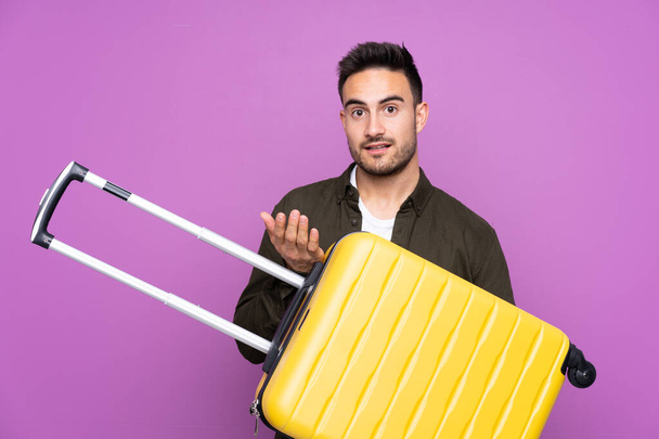 Mladý pohledný muž přes izolované fialové pozadí v dovolené s cestovní kufr - Fotografie, Obrázek