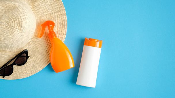 Blank sunblock spray package and sunscreen lotion bottle on blue background with sunglasses and beach hat. Top view with copy space. Sun protection cosmetic products, summer skin care concept - Foto, Imagen