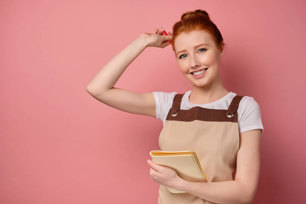 A cute red-haired girl with a high bun in a beige apron is smiling, holding a notebook to herself and putting a pencil behind ear - Фото, зображення
