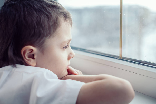 one sad little boy sitting near the window at the day time. Concept of sorrow. - Фото, изображение