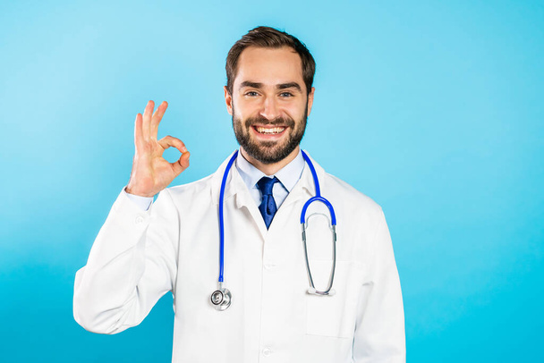 Portrait of smiling positive doctor in professional medical white coat showing OK sign gesture. Doc isolated on blue background. - Foto, afbeelding