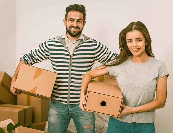 Young couple smiling and holding boxes while standing among unpacked boxes - Photo, image