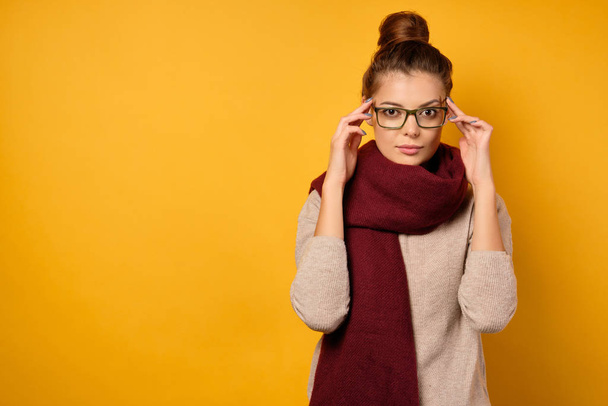 A beautiful brunette with a bun on head and glasses stands on a yellow background in a sweater and a scarf, looks at the camera - Foto, immagini