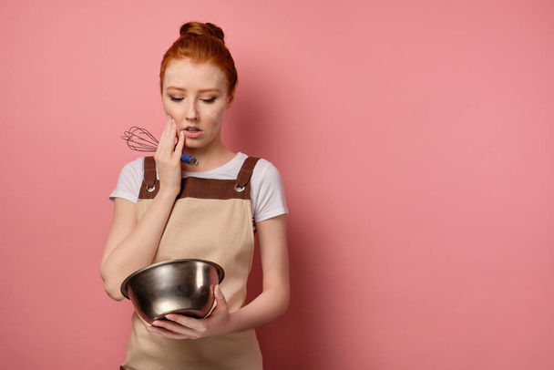 A cute red-haired girl with a high bun in a apron and a face in flour stands on a pink background and looks doubtfully into a bowl - Foto, afbeelding
