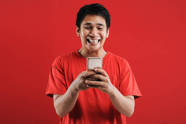 Boldog ázsiai férfi olvas új szöveget mobil, elszigetelt piros háttér - Fotó, kép