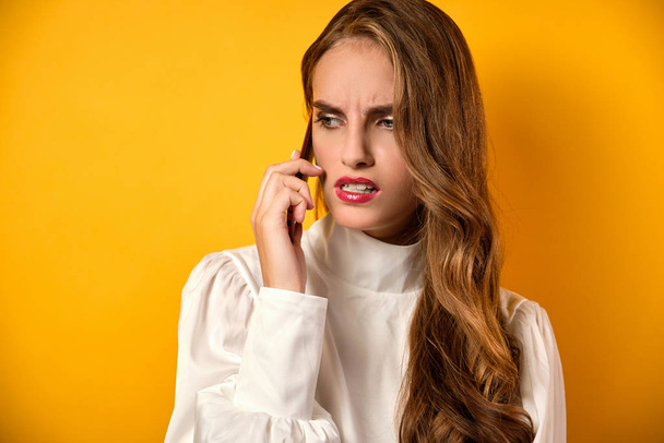 A beautiful girl with clean skin and red lipstick stands on a yellow background and speaks annoyingly on the phone. - Foto, imagen