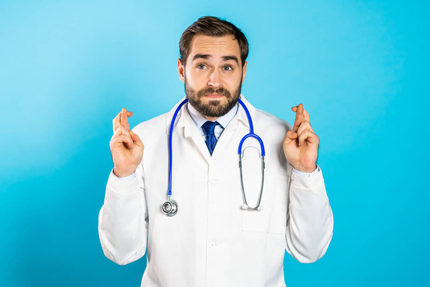 Handsome doctor man in white professional coat praying over blue background. European race guy begging someone. - Фото, изображение