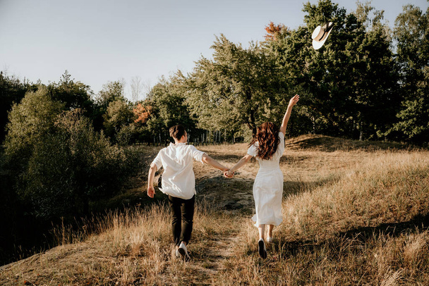 Loving couple walks and runs at sunset in the summer on the nature. Brunette girl in a hat and a light dress. Love story. - Fotoğraf, Görsel