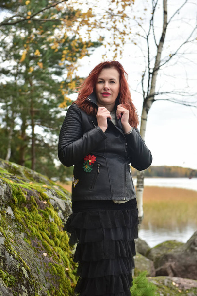 red-haired woman in a black leather jacket and skirt in an oxer park among stones - Photo, Image