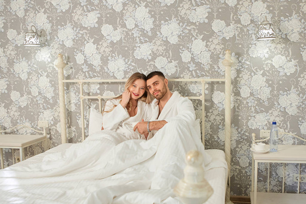 Felice giovane coppia in una camera d'albergo su un grande letto matrimoniale in accappatoi bianchi
 - Foto, immagini