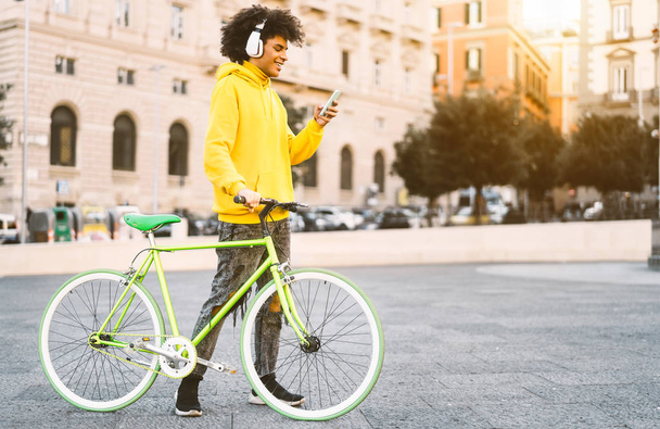 Glücklicher Afro-Mann mit Handy im Freien - Junger Mann hat Spaß mit Kopfhörern Musik zu hören, während er mit dem Fahrrad in der Stadt unterwegs ist - Lifestyle- und Technologiekonzept der Millennial Generation - Foto, Bild