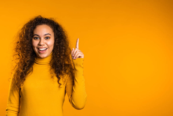 Portrait of young thinking pondering woman having idea moment pointing finger up on yellow studio background. Smiling happy girl showing eureka gesture. - Photo, image
