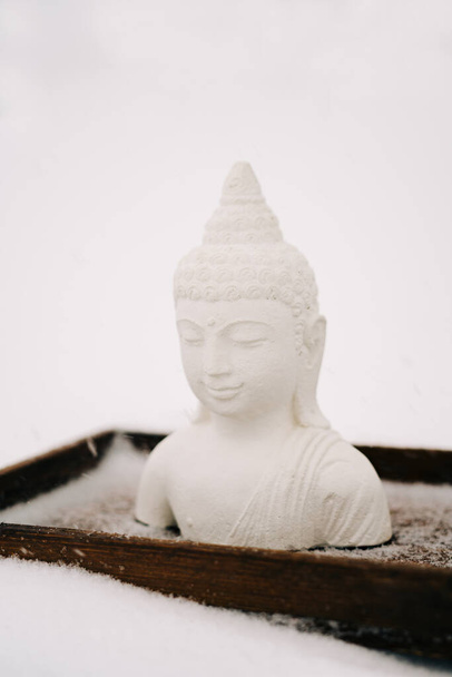 Figura de Buda blanco hecha de yeso en un soporte de madera durante una nevada
 - Foto, Imagen