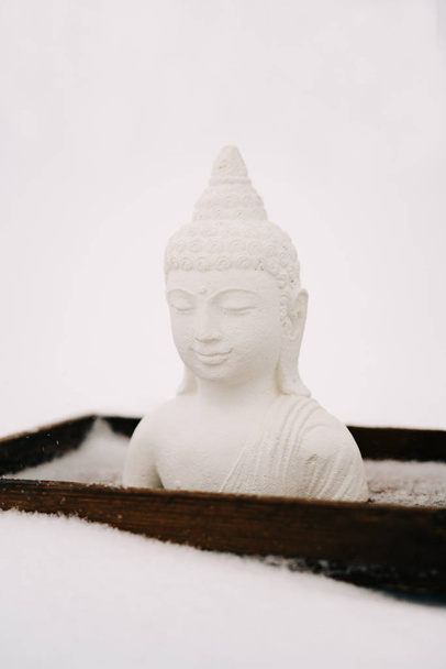 Figura de Buda blanco hecha de yeso en un soporte de madera durante una nevada
 - Foto, Imagen