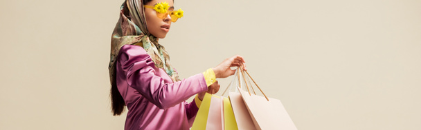 panoramic shot of trendy african american girl in sunglasses with flowers and head scarf holding shopping bags isolated on beige  - Photo, Image