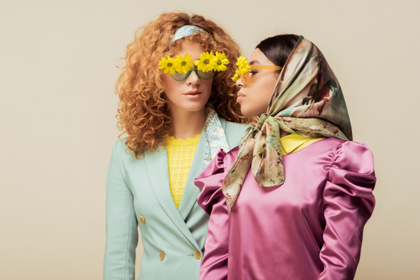 trendy african american girl and curly redhead woman in sunglasses with flowers posing isolated on beige - Fotografie, Obrázek