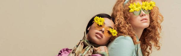 панорамний знімок модної афроамериканської дівчини і рудої жінки в сонцезахисних окулярах з квітами позує ізольовано на бежевому
 - Фото, зображення