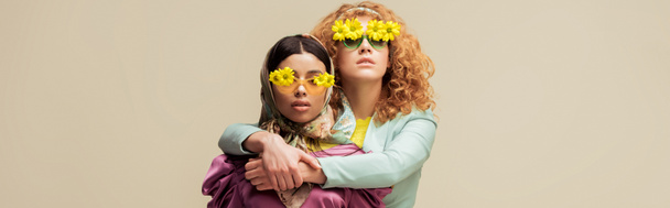 panoramatický záběr atraktivní africké americké dívky a kudrnaté zrzky žena v slunečních brýlích s květinami pózovat izolované na béžové - Fotografie, Obrázek