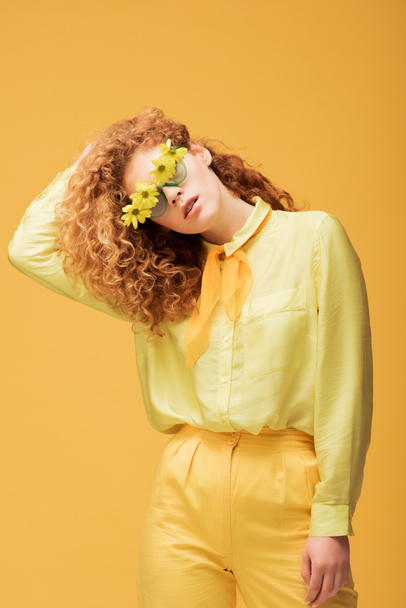 стильна руда жінка в сонцезахисних окулярах з квітами позує ізольовано на жовтому
  - Фото, зображення