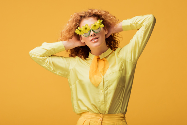 μοντέρνα κοκκινομάλλα γυναίκα με γυαλιά ηλίου με λουλούδια που ποζάρουν απομονωμένα σε κίτρινο  - Φωτογραφία, εικόνα