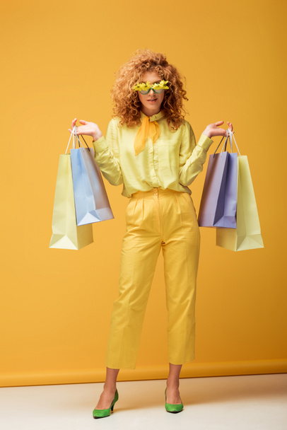 stylish redhead woman in sunglasses with flowers holding shopping bags on yellow  - Photo, Image