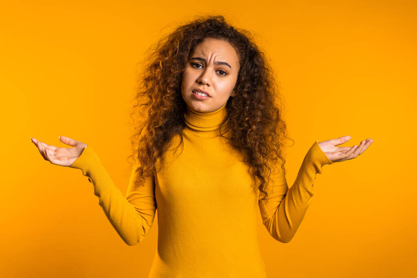  Young curly unsure girl shrugs her arms, makes gesture of I dont know, care, cant help anything .Young mixed race woman on yellow background - Foto, imagen