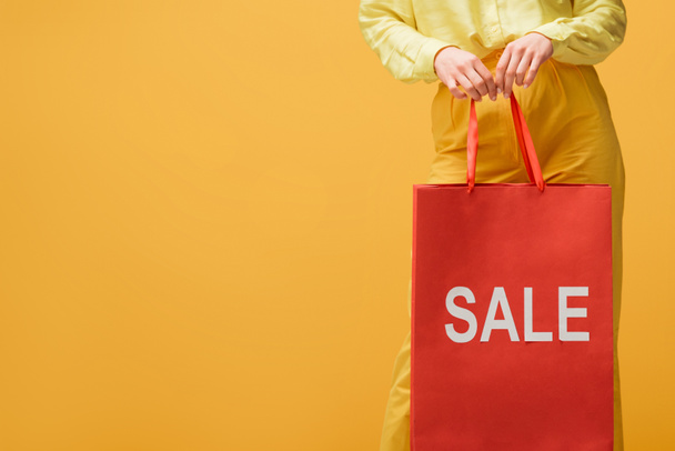 cropped view of young woman holding shopping bag with sale lettering while standing isolated on yellow  - Zdjęcie, obraz