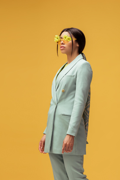 stylish african american girl in suit and sunglasses with flowers posing isolated on yellow  - Foto, Bild