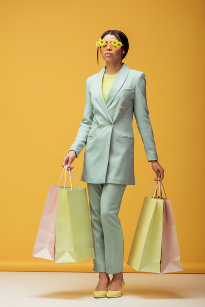stylish african american girl in suit and sunglasses with flowers holding shopping bags on yellow  - Фото, изображение