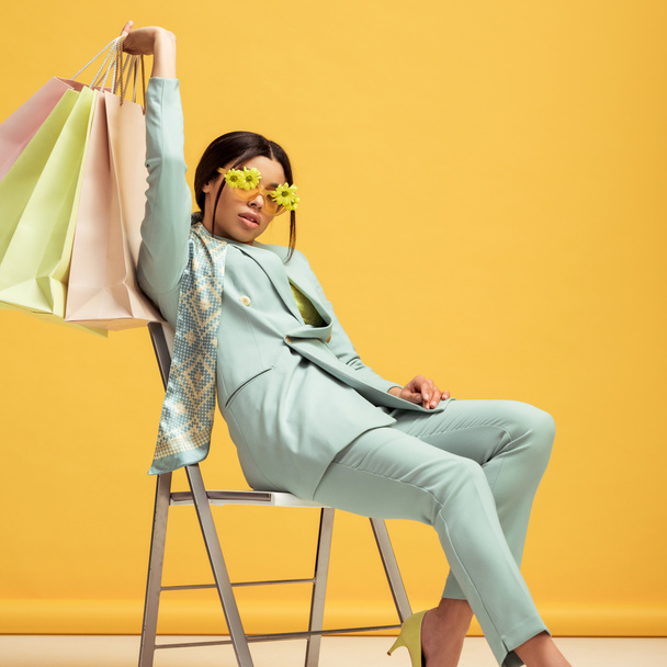 κομψή Αφροαμερικανίδα με κοστούμι και γυαλιά ηλίου με λουλούδια που κρατούν τσάντες για ψώνια ενώ κάθεται σε κίτρινο  - Φωτογραφία, εικόνα