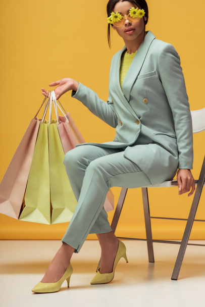 trendy african american woman in suit and sunglasses with flowers holding shopping bags while sitting on yellow  - Foto, immagini