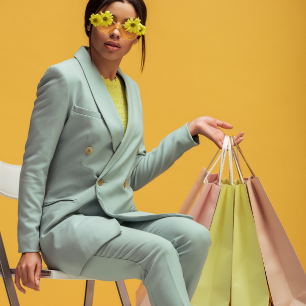 stylish african american woman in suit and sunglasses with flowers holding shopping bags while sitting isolated on yellow  - Фото, изображение