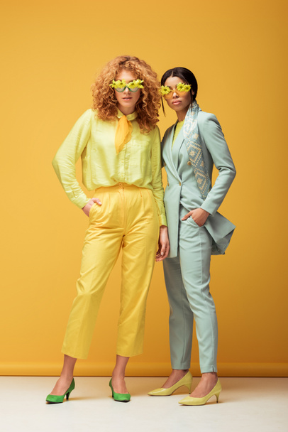 attractive multicultural women in sunglasses with flowers standing with hands in pockets on yellow  - Photo, Image