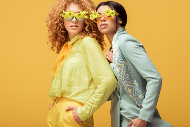 attractive multicultural women in sunglasses with flowers standing with hands in pockets isolated on yellow  - Фото, зображення